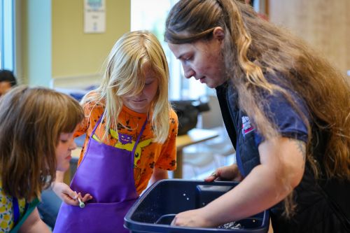 A young boy in an orange shirt and purple apron looks into a blue bin as an adult helps them with an activity.